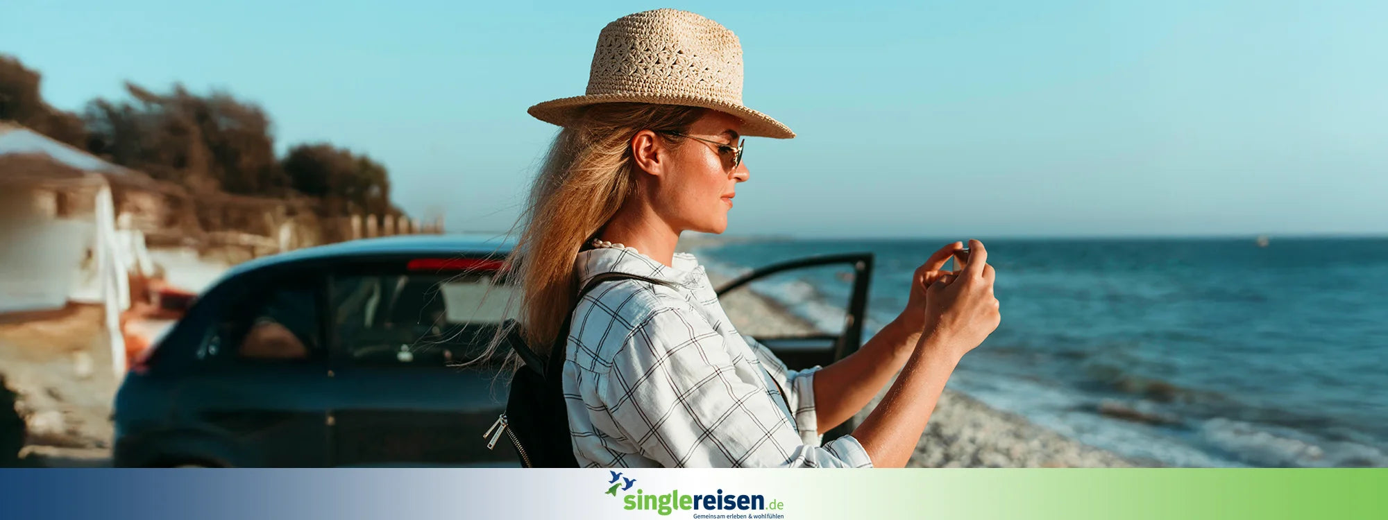 Eine junge Frau mit Strohhut und Sonnenbrille fotografiert im Sommerurlaub mit ihrem Smartphone das Meer neben einem geparkten Auto am Strand – singlereisen.de.