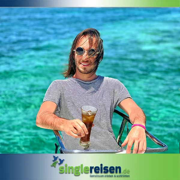 Ein lächelnder junger Mann mit Sonnenbrille sitzt mit einem Kaltgetränk an Bord eines Bootes vor türkisblauem Meer – Singlereisen ab 30 Jahre auf singlereisen.de.