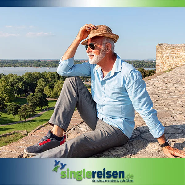 Ein eleganter älterer Mann mit grauem Bart, Sonnenbrille und Strohhut sitzt entspannt auf einer alten Steinmauer und blickt in die Ferne – Singlereisen ab 50 Jahre auf singlereisen.de.