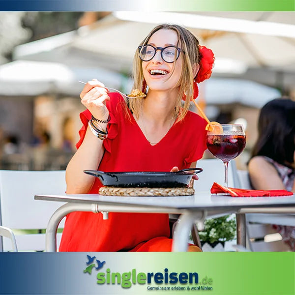 Eine lachende Frau im roten Kleid mit Brille isst Paella und genießt ein Glas Sangria in einem Außencafé – Kultur & Genuss auf singlereisen.de.