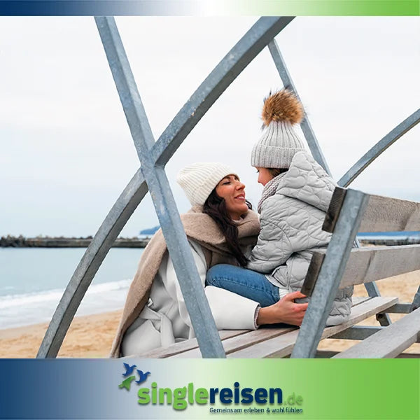 Eine Mutter und ihre Tochter in warmer Winterkleidung mit Mützen sitzen eng aneinander gekuschelt auf einer Holzbank am Strand – Singlereisen mit Kind auf singlereisen.de.