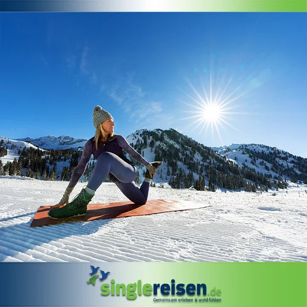 Eine Frau in Sportkleidung und Mütze dehnt sich im Schnee auf einer Matte vor einer verschneiten Bergkulisse im strahlenden Sonnenschein – Ski • Snowboard • Action auf singlereisen.de.