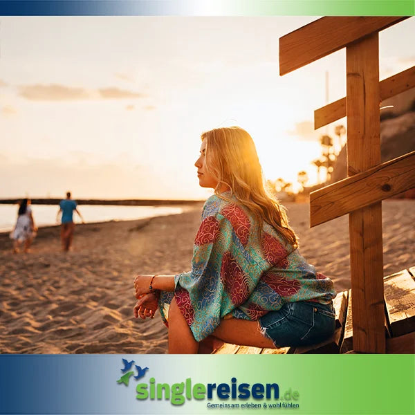Eine junge Frau im bunten Strand-Poncho blickt bei Sonnenuntergang am Sandstrand aufs Meer hinaus – Sonne • Strand • Meer auf singlereisen.de.