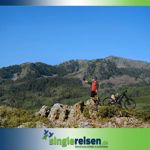 Eine Person mit Fahrradhelm steht mit einem E-Mountainbike auf einem Felshügel vor einer bewaldeten Bergkulisse unter blauem Himmel – Wanderreisen • Radfahren auf singlereisen.de.